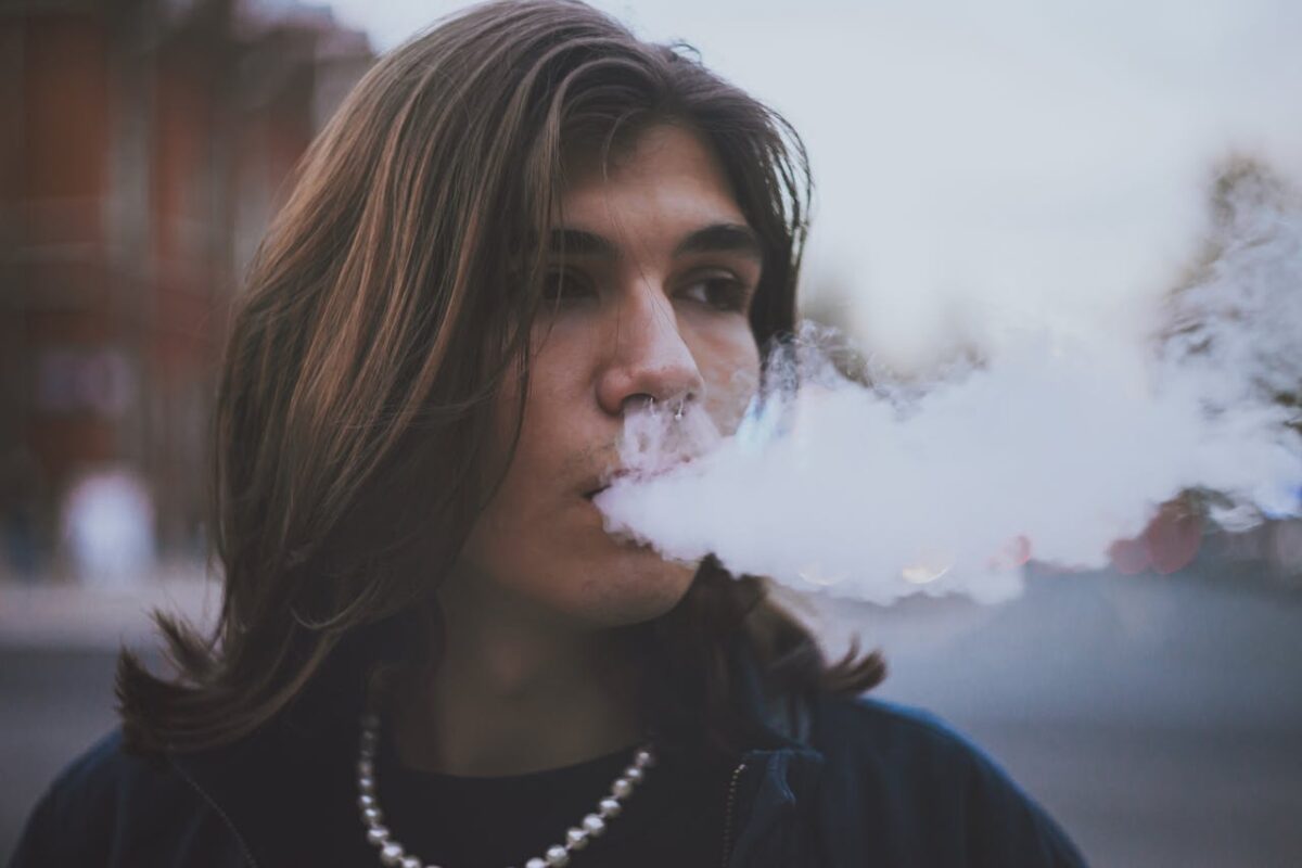 our-experience Stylish young man with long hair vaping outdoors, showcasing modern lifestyle.