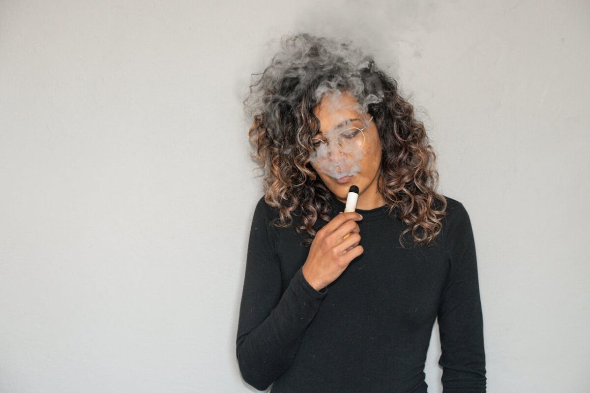 about-img Portrait of a woman with curly hair vaping indoors, featuring smoke and a relaxed expression.