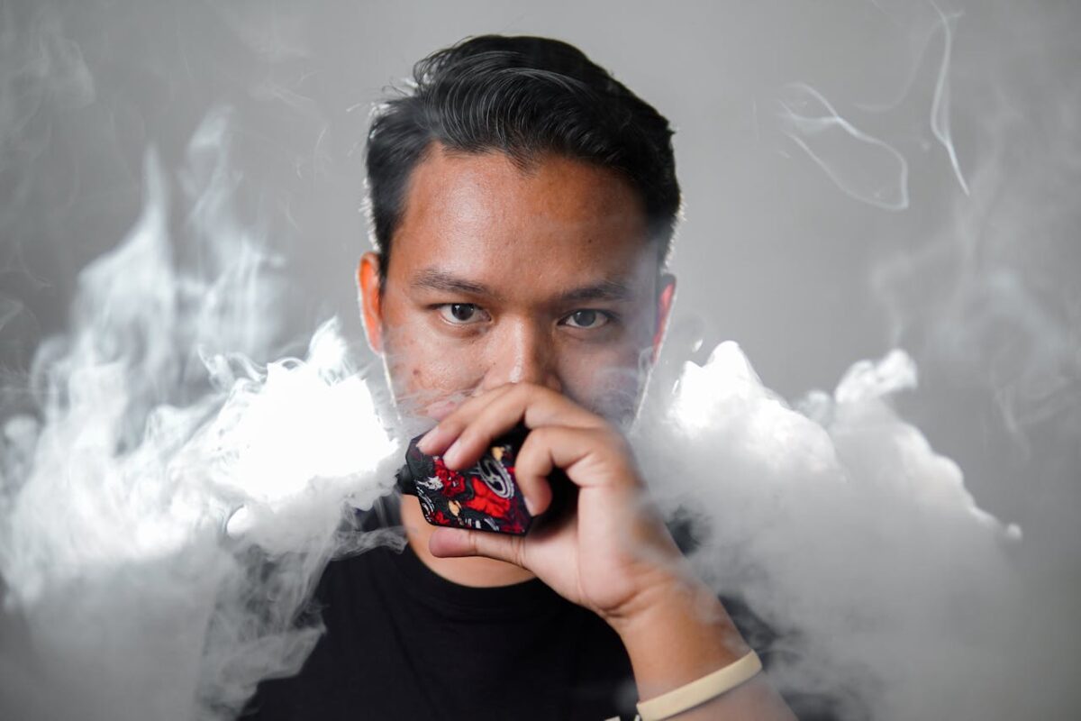 services-03 Portrait of a young man with black hair vaping, surrounded by smoke against a gray background.