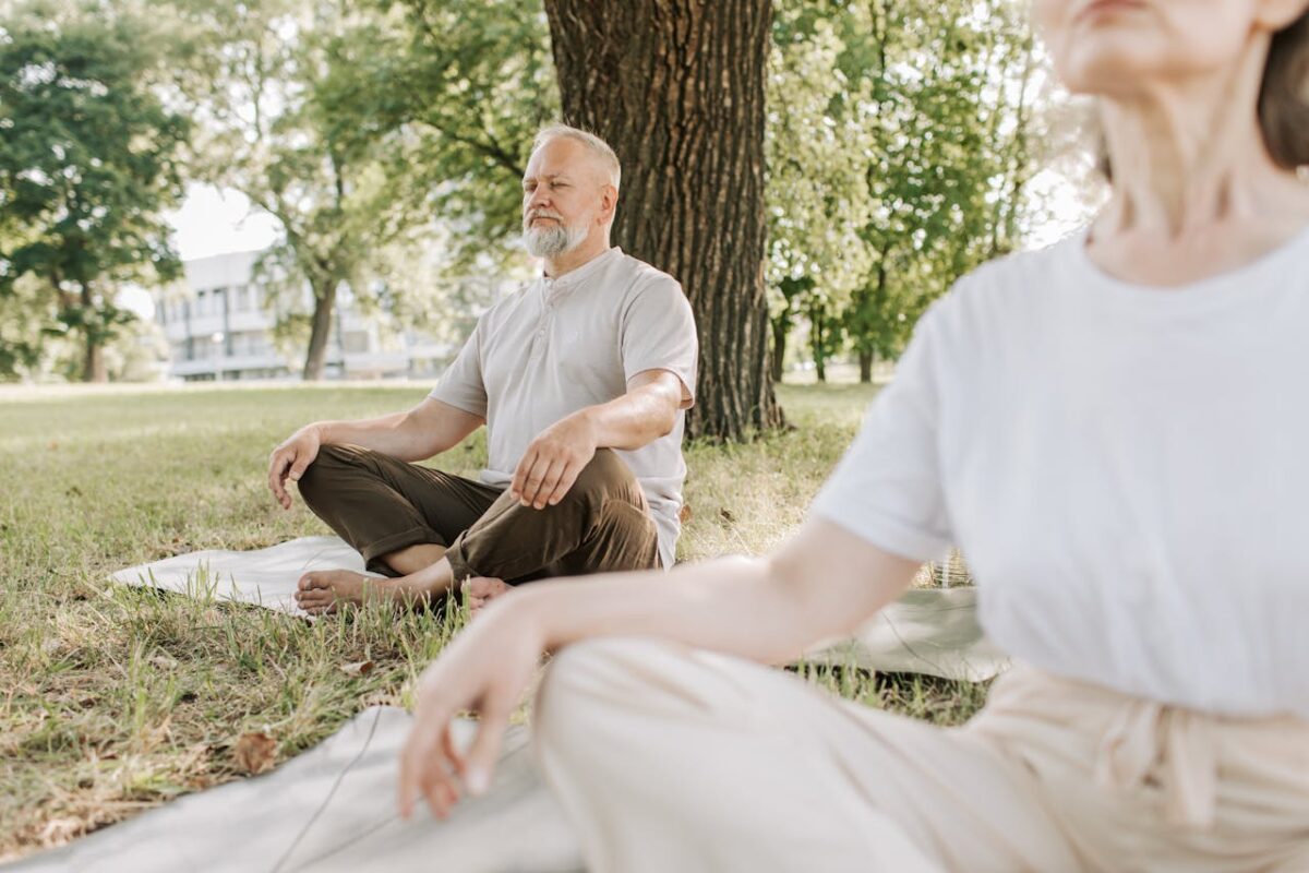 services-02 Elderly man and woman meditating outdoors on yoga mats in a peaceful park setting.